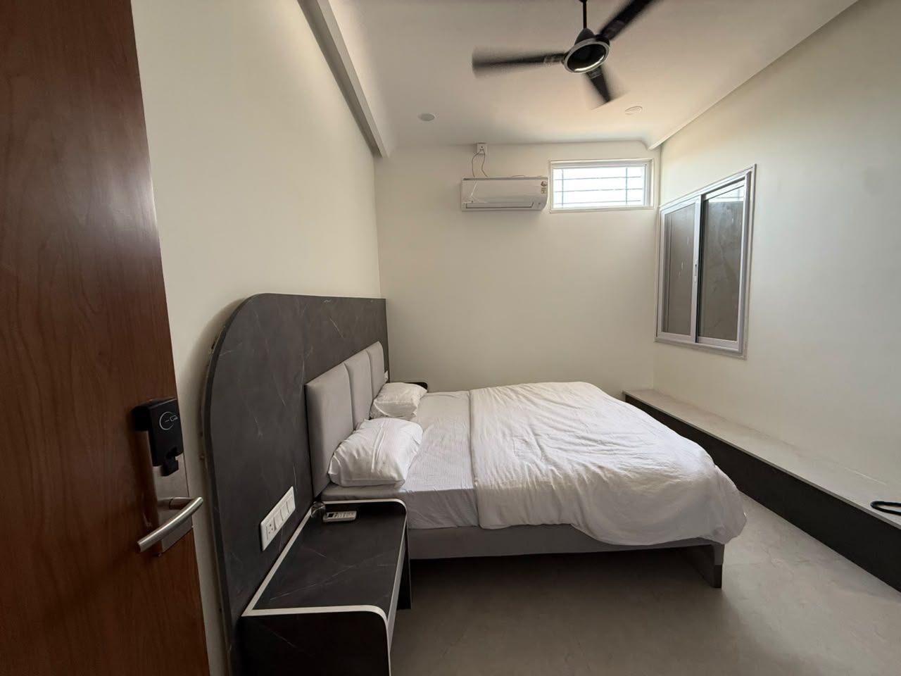 A bedroom with a bed, a bedside table, a ceiling fan, an air conditioning unit, and a window.