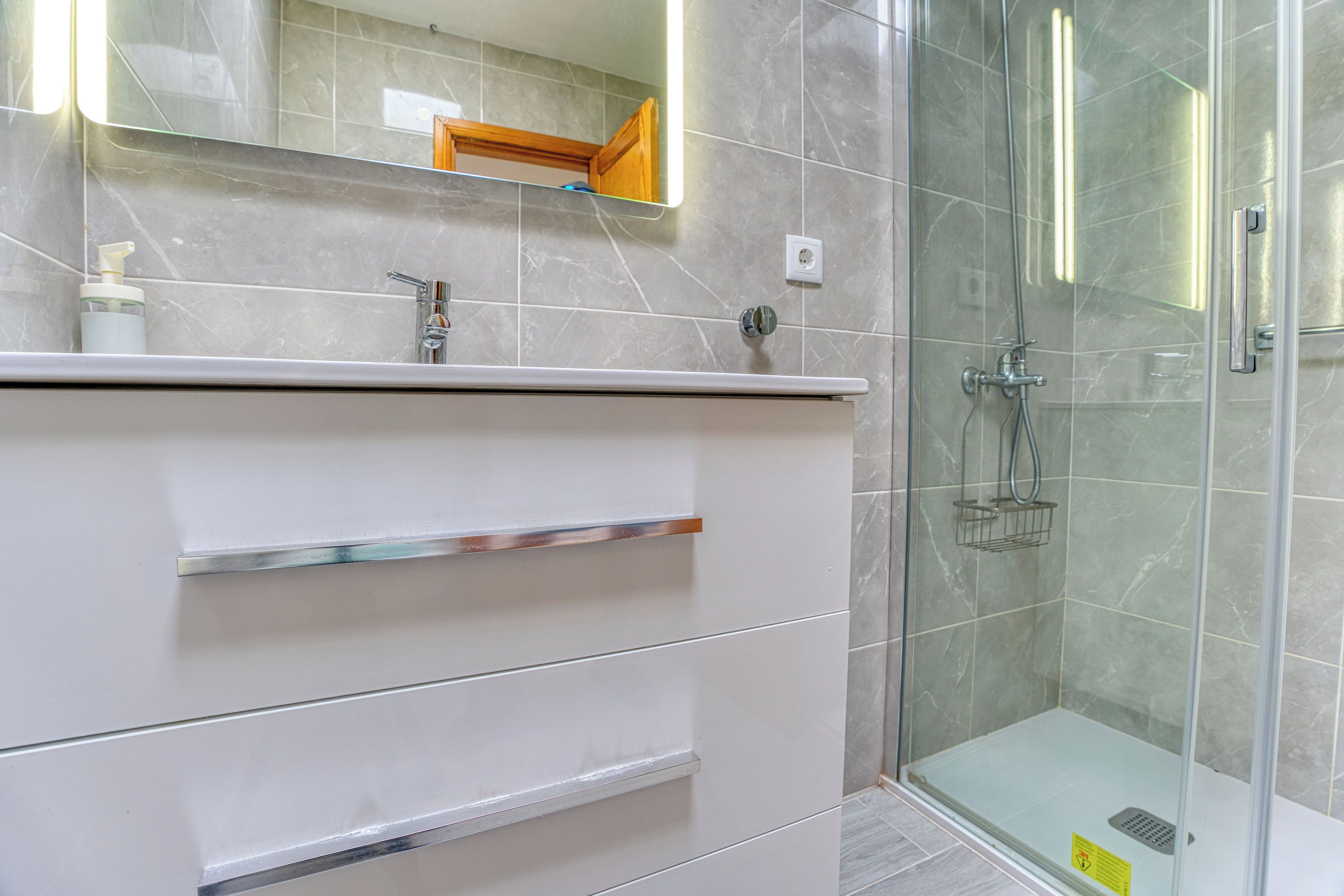 Un baño con cabina de ducha de vidrio, tocador blanco con cajones y espejo sobre el tocador.