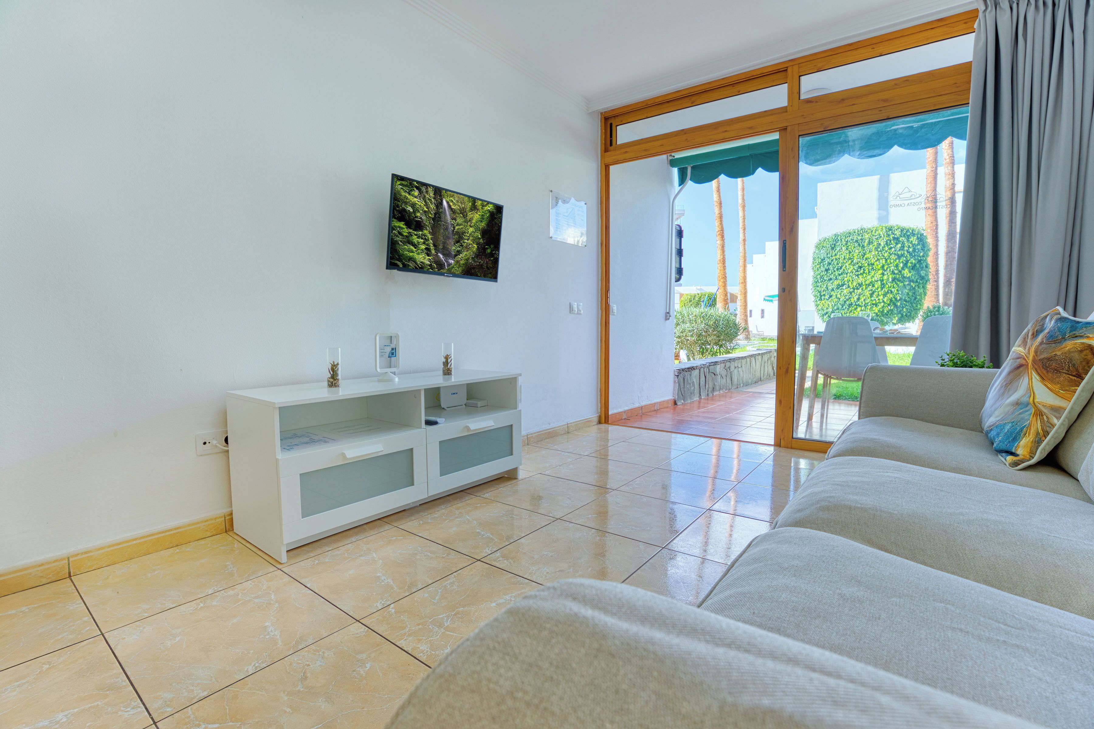 A modern living room with a white sofa, a flat-screen TV mounted on the wall, and a sliding glass door leading to an outdoor area.