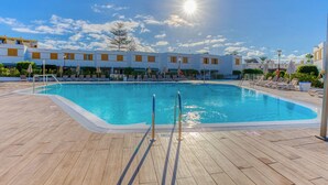 A swimming pool with sun reflecting on the water, surrounded by lounge chairs and a building with a row of windows. - Las Brisas (San Bartolomé de Tirajana)