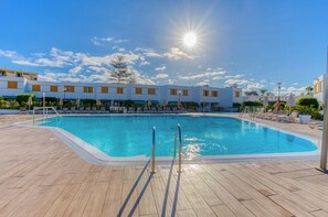 Une piscine dont l’eau scintille sous les rayons du soleil, entourée de chaises longues et bordée d’un bâtiment dont les fenêtres s’alignent en rangée.