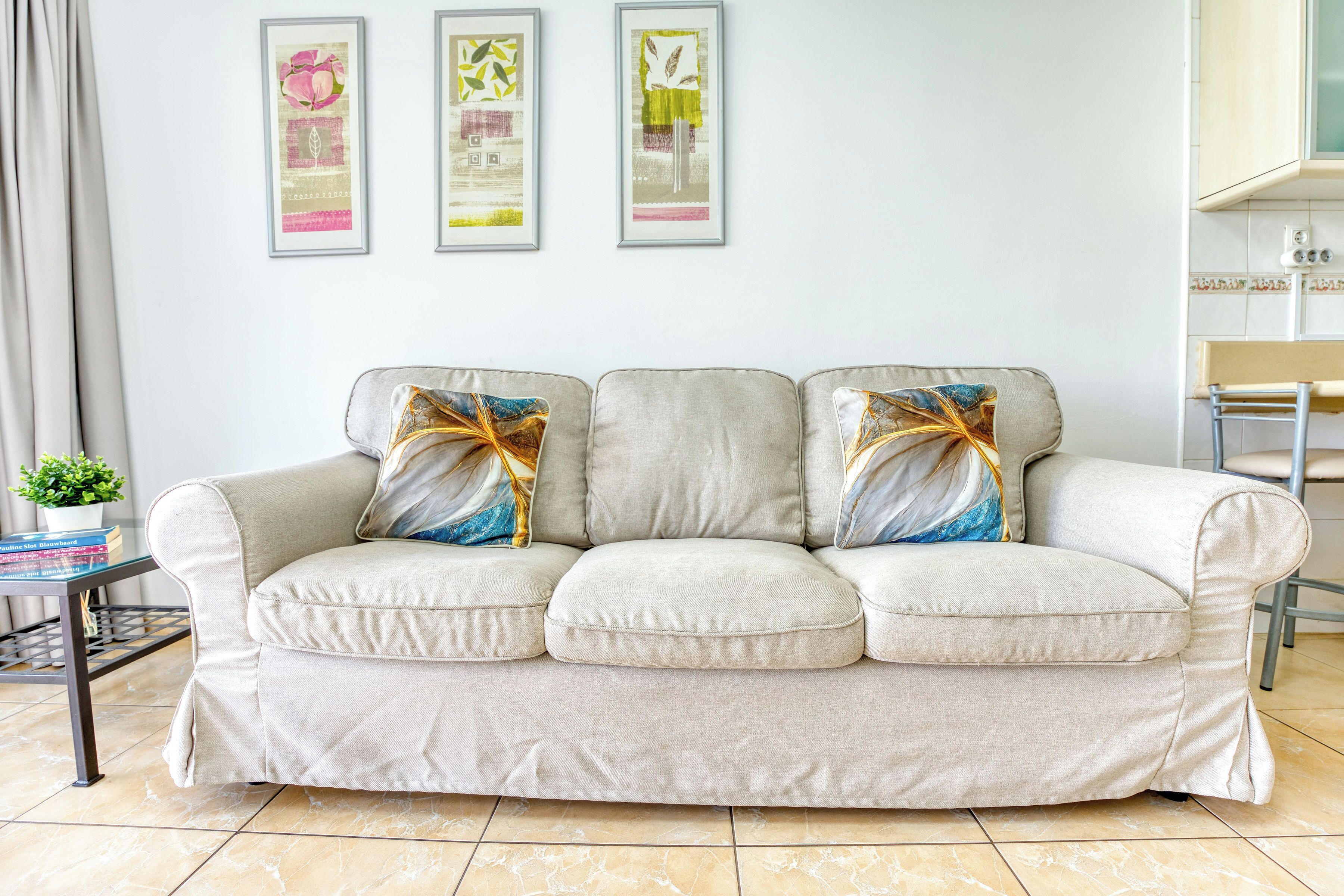 Una sala de estar con un sofá color beige, dos almohadones decorativos, una mesita de noche con una planta y tres obras de arte enmarcadas en la pared.