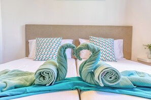 Una cama con cabecera, dos almohadas estampadas y toallas dispuestas en forma de corazón.