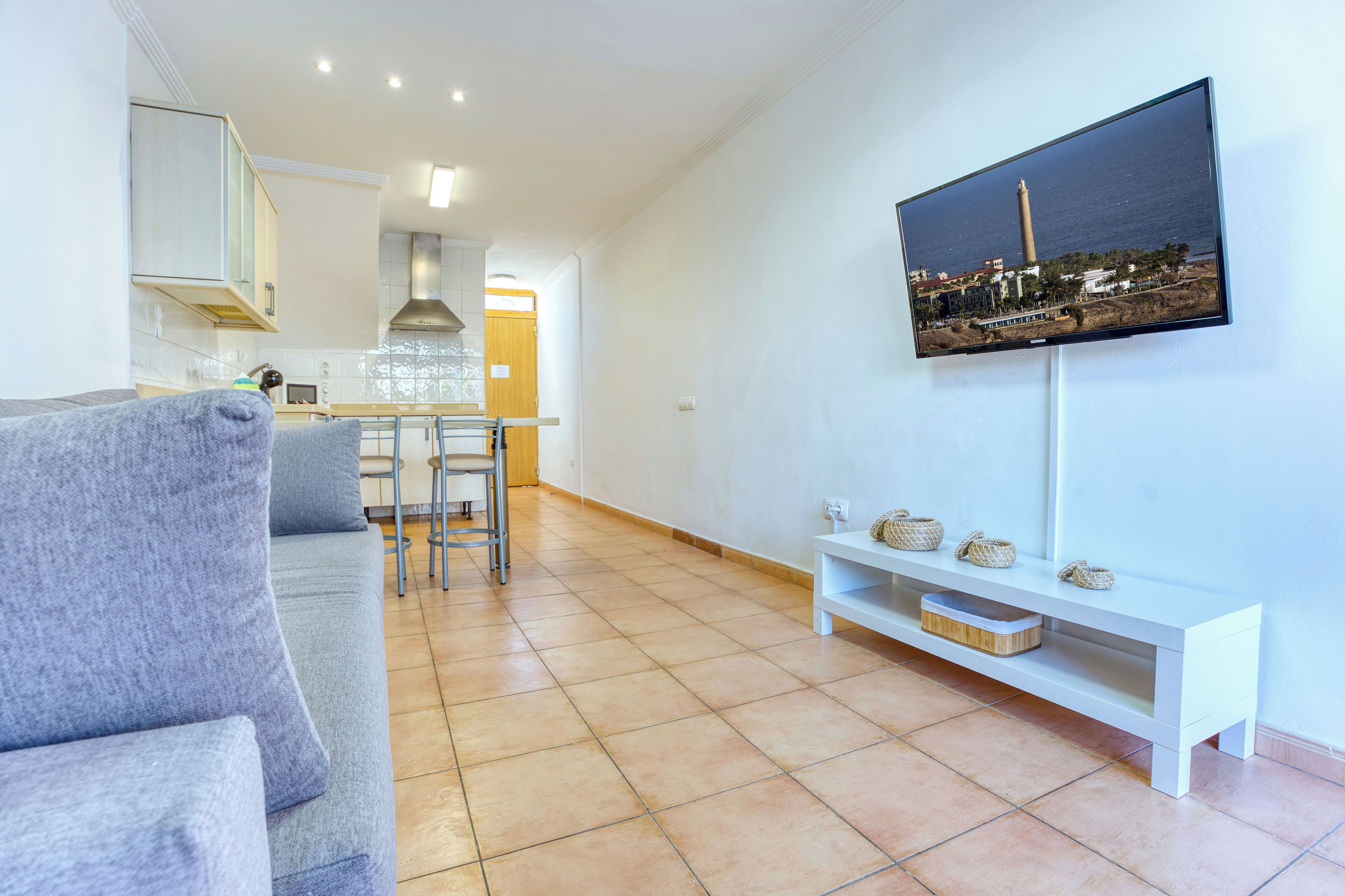 A modern living room with a grey sofa, a TV mounted on the wall, and a kitchen in the background.
