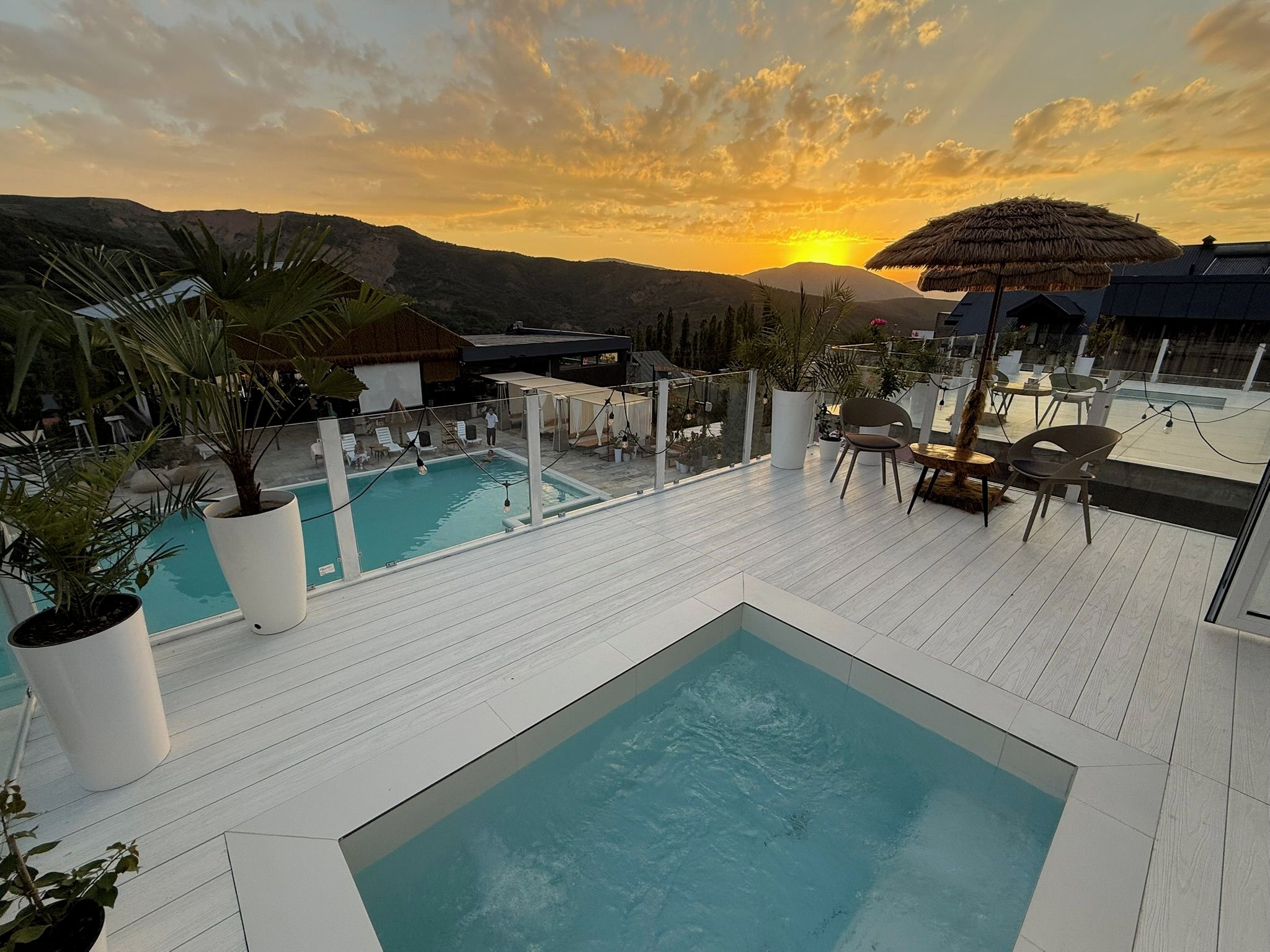 A rooftop pool area with multiple pools and lounge chairs, surrounded by potted plants and overlooking a mountainous landscape.