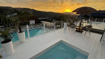 A rooftop pool area with multiple pools and lounge chairs, surrounded by potted plants and overlooking a mountainous landscape.