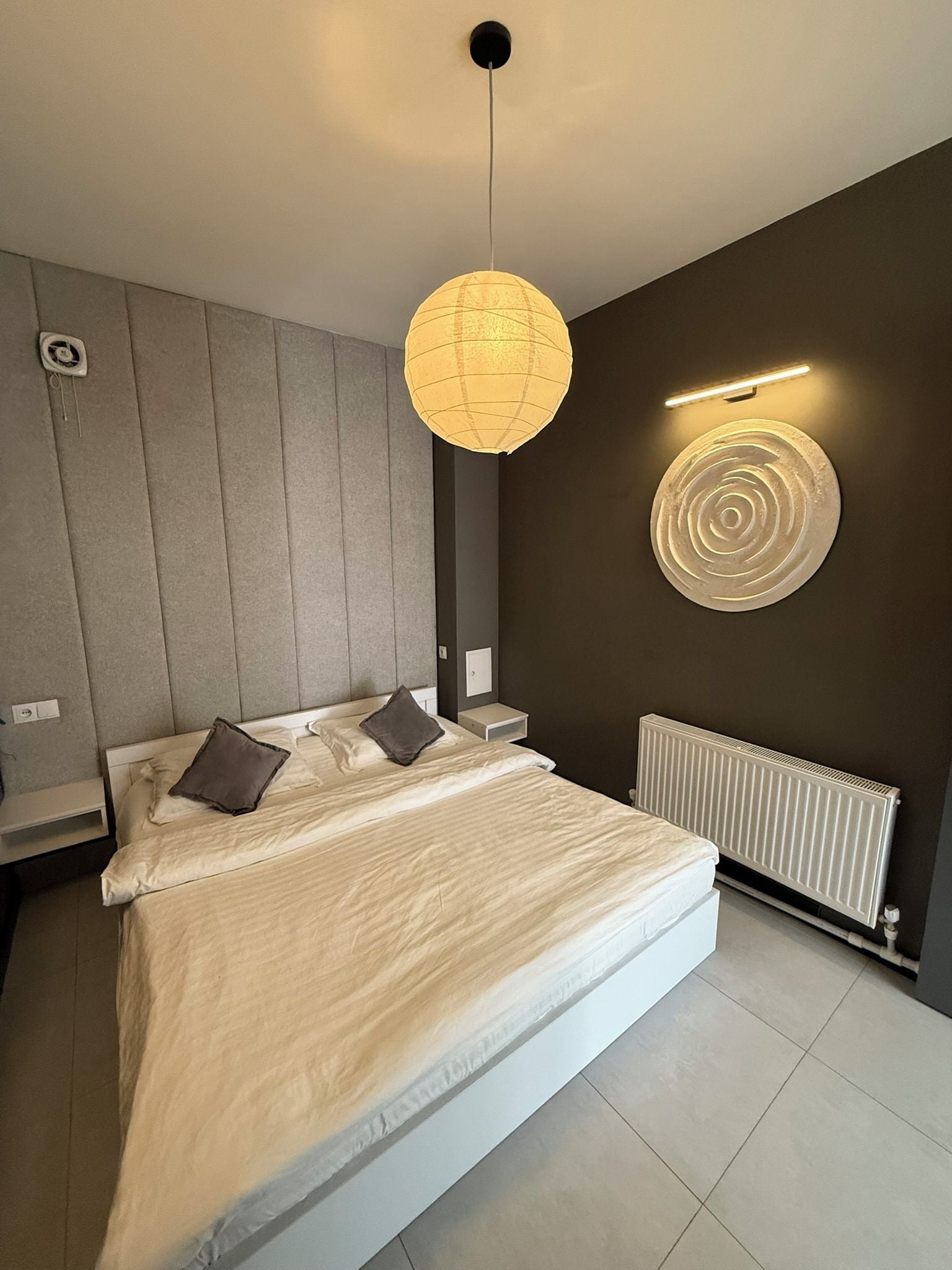 Un dormitorio moderno con una cama grande, una lámpara colgante, una luz de pared y un radiador.