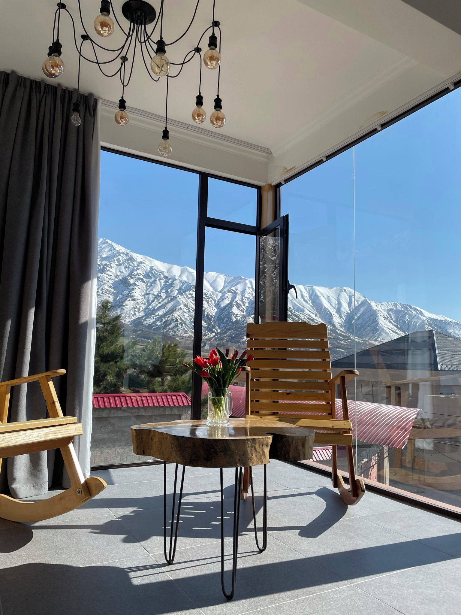 A balcony with a wooden table, chairs, and a vase of flowers. A mountain view is visible through the large windows.