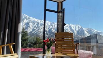 A balcony with a wooden table, chairs, and a vase of flowers. A mountain view is visible through the large windows.