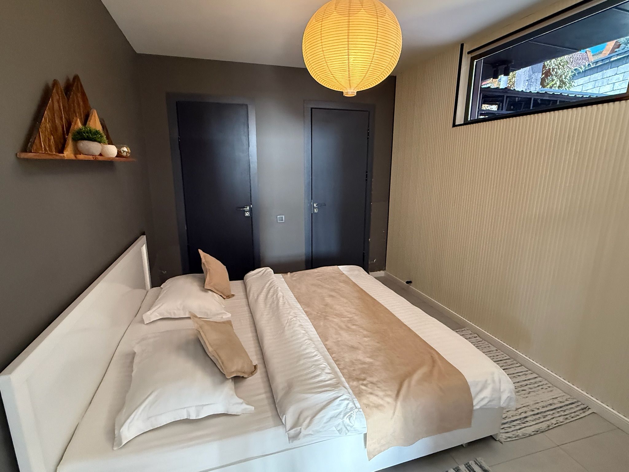 Quarto com cama, duas portas, prateleira com objetos decorativos e luminária pendente.