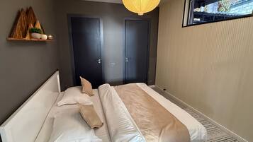 Quarto com cama, duas portas, prateleira com objetos decorativos e luminária pendente.