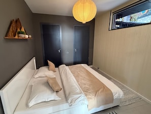 A bedroom with a bed, two doors, a shelf with decorative items, and a hanging light fixture.