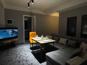 A modern living room with a flat-screen TV, a dining table set for two, a sofa, and a bright orange chair.