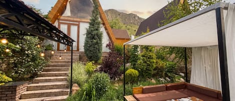 Un espacio de estar moderno al aire libre con una mesa y bancos de madera, rodeado de abundante vegetación y un telón de fondo montañoso.