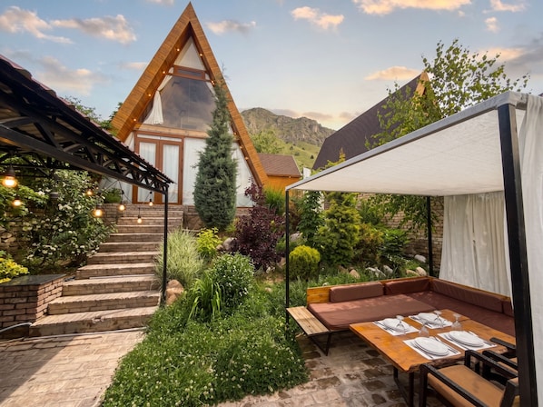 A modern outdoor seating area with a wooden table and benches, surrounded by lush greenery and a mountain backdrop.