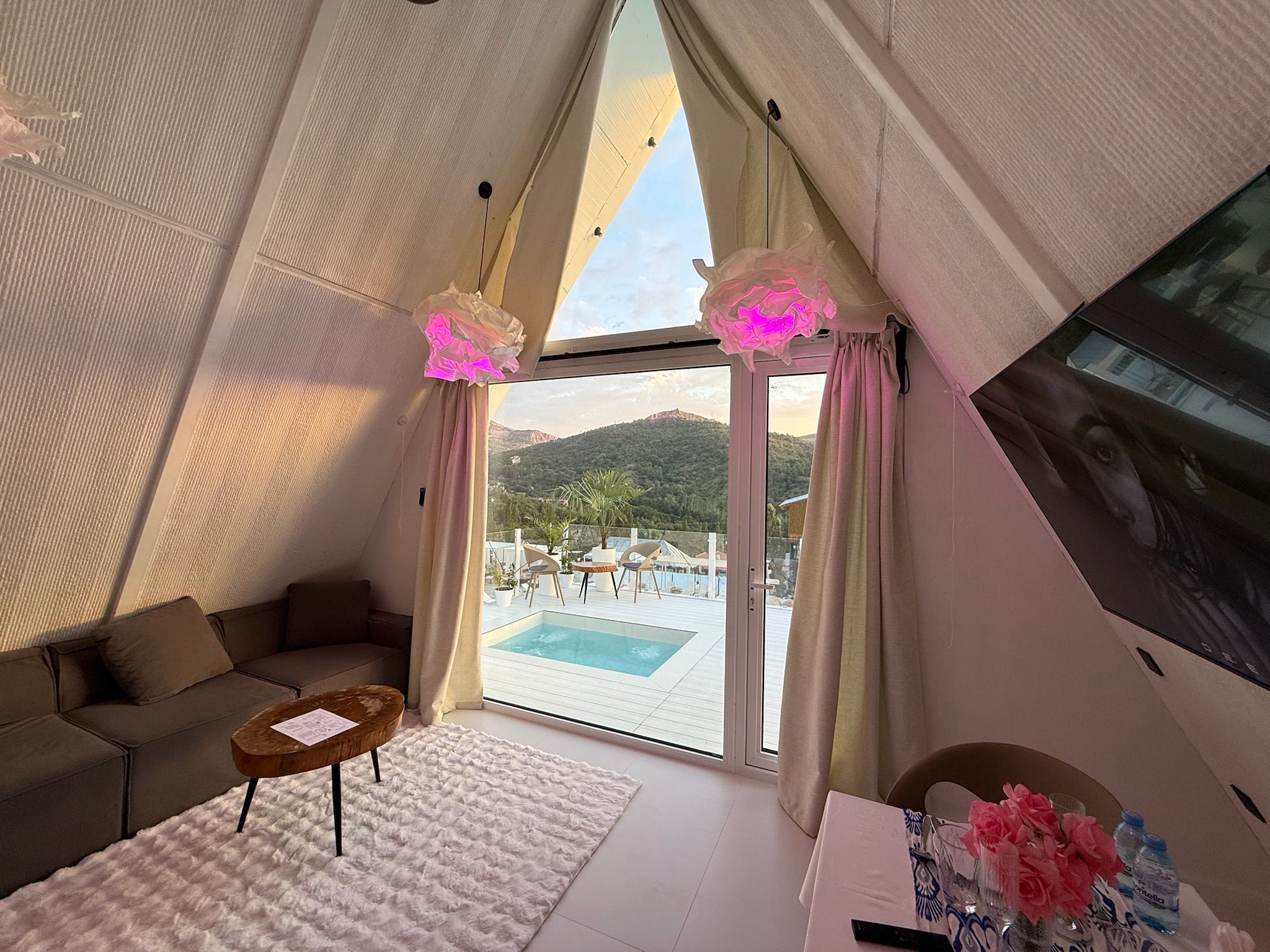 Una habitación acogedora con sofá, una mesita, una alfombra blanca y vistas a una piscina y montañas a través de un ventanal amplio.