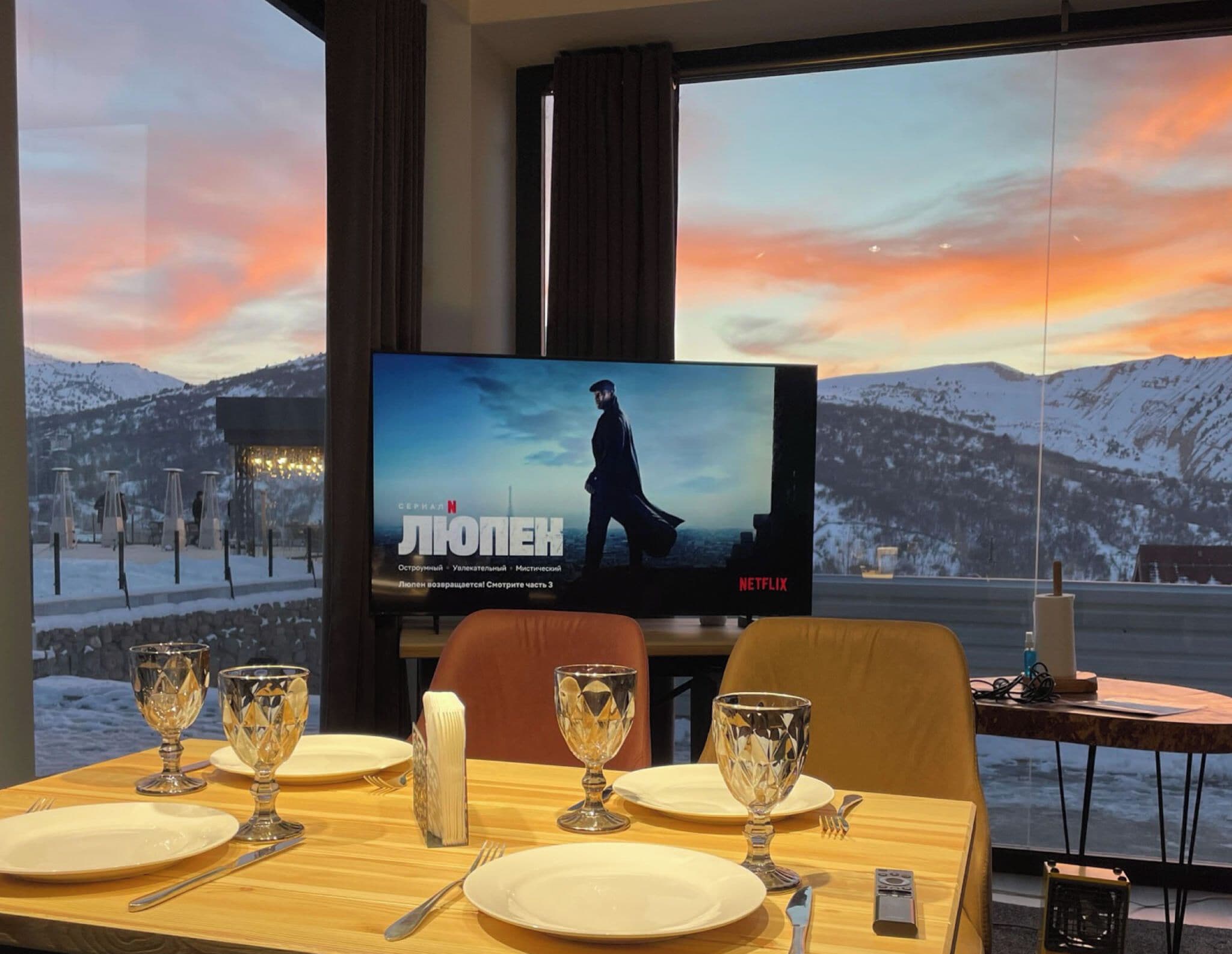 A hotel room with a large window offering a scenic view of snow-covered mountains and a sunset. A flat-screen TV is mounted on the wall, displaying a Netflix show. The room is furnished with a wooden dining table set for two, complete with plates, glasses, and cutlery.