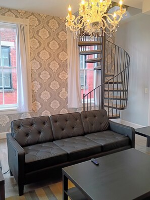 A living room with a black leather sofa, a black coffee table, a chandelier, and a spiral staircase. - Edge of Downtown Cincinnati Two Floor Condo (Cincinnati)
