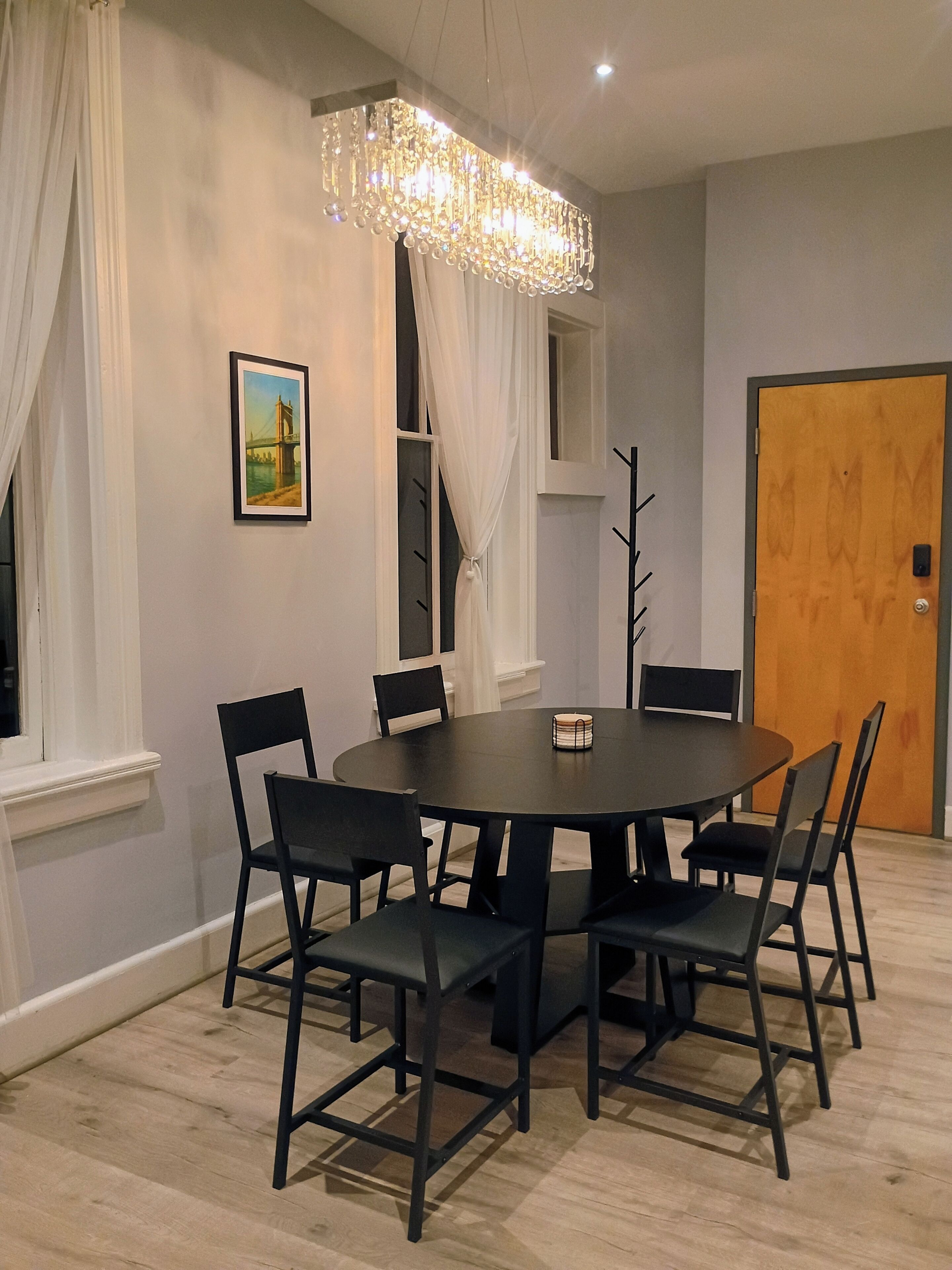 A dining area with a round table, six chairs, a chandelier, a painting, and a door.
