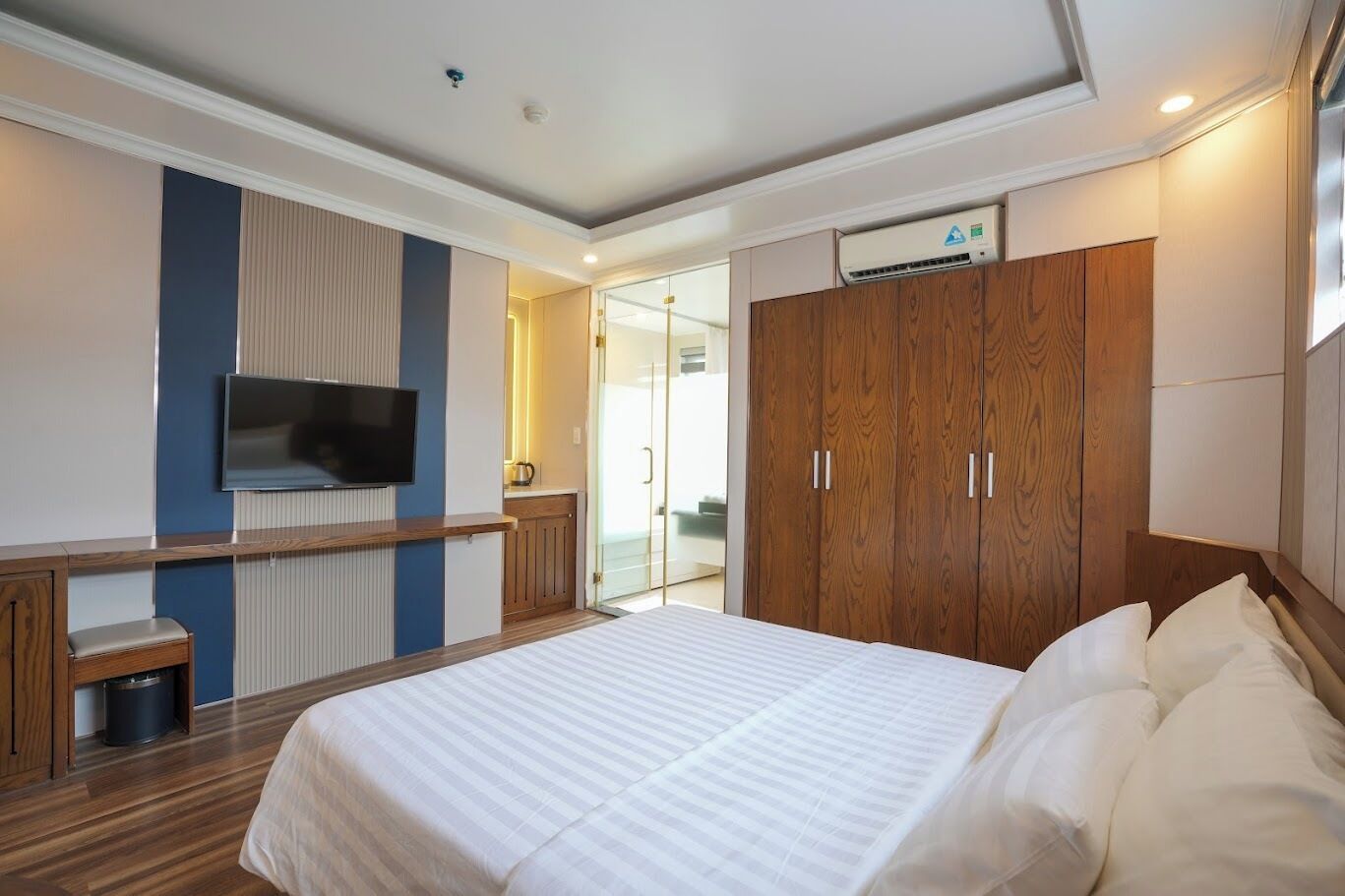 A hotel room with a bed, a TV mounted on the wall, a wardrobe, and a small seating area with a bench and trash bin.