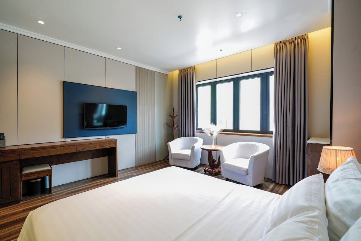 Quarto de hotel com uma cama grande, televisão na parede, secretária, duas cadeiras e janela com cortinas.