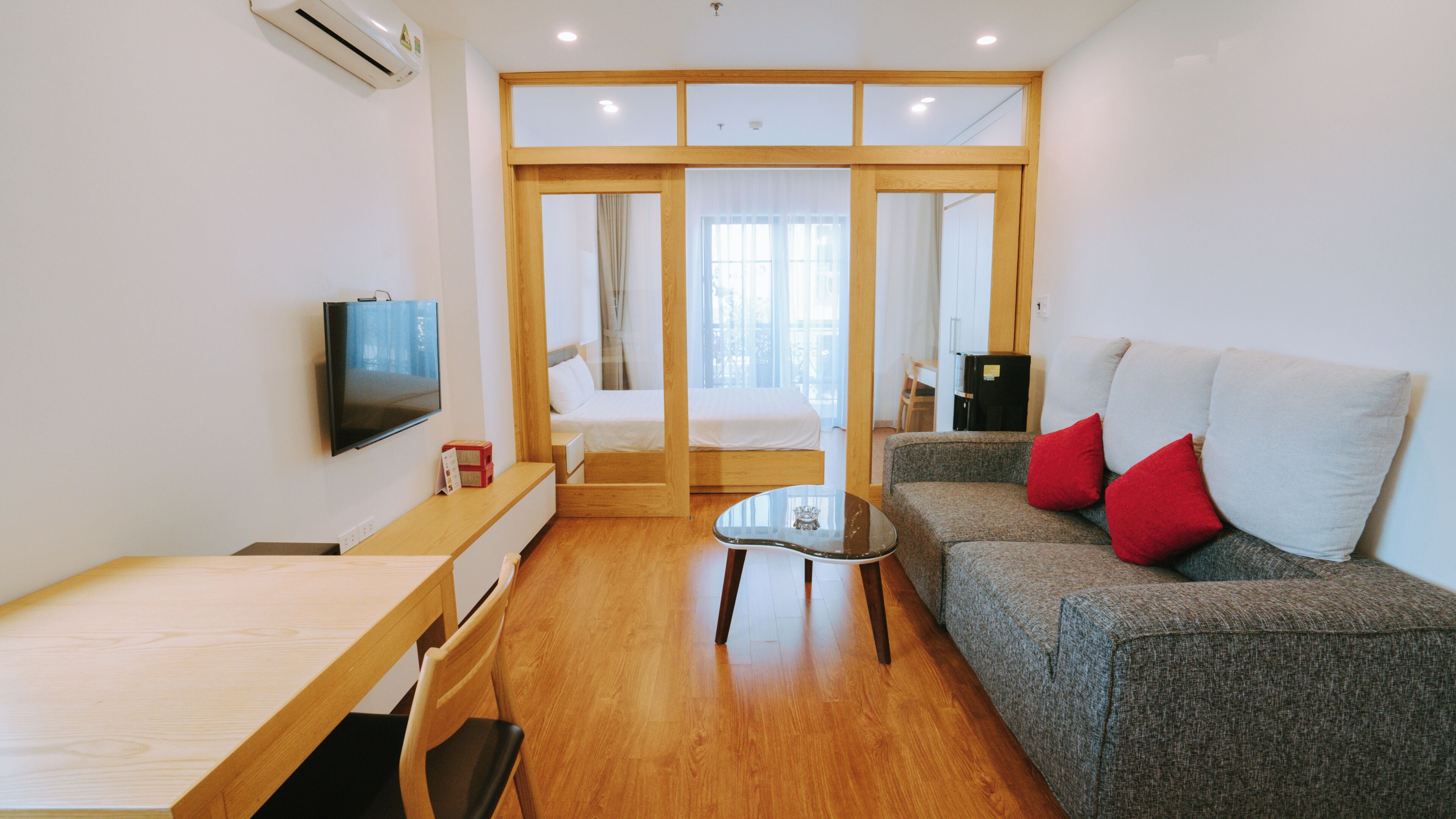 A modern room with a bed, a sofa with red cushions, a TV, a wooden desk, and a glass coffee table.