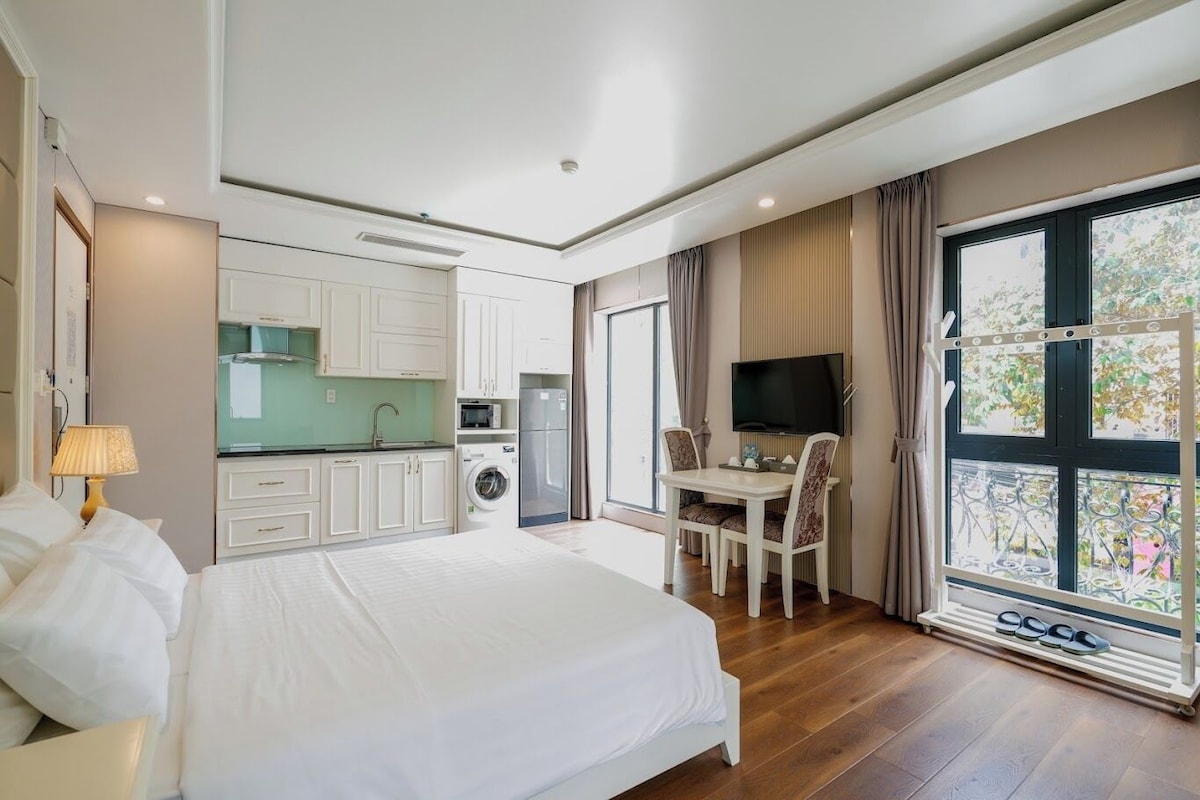 Quarto de hotel com cama, escrivaninha, televisão, máquina de lavar roupa e uma janela grande com vista.