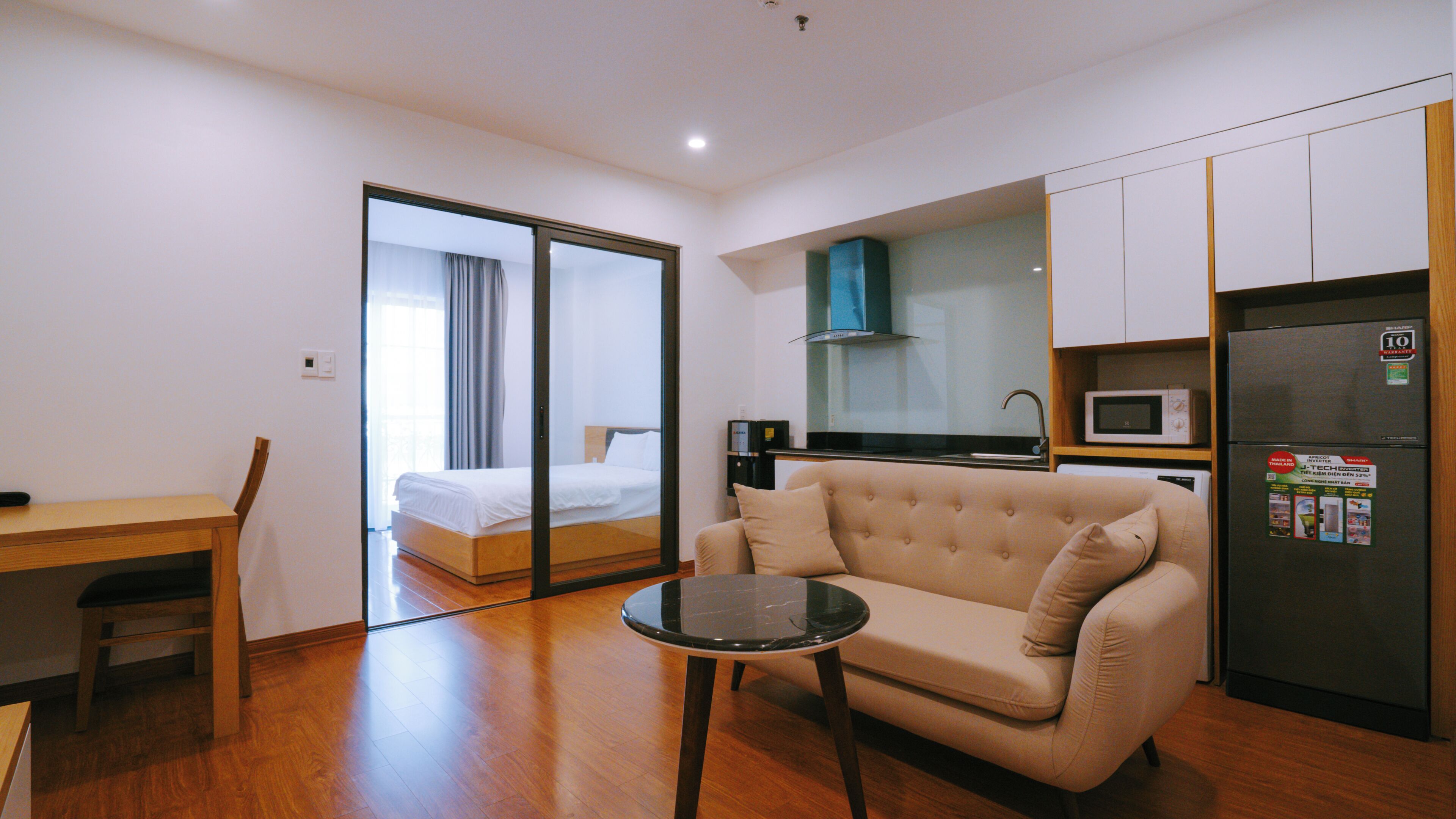 A compact hotel room with a bed, a sofa, a small table, a kitchenette, and a sliding door leading to a balcony.