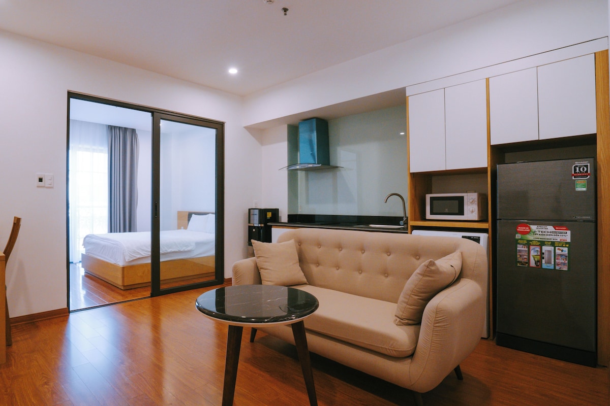 Quarto de hotel compacto com cama, sofá, mesinha, cozinha compacta e porta de correr para uma varanda.