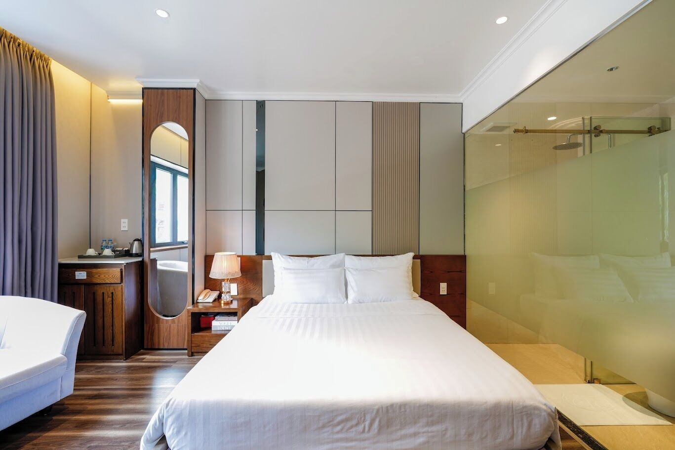 Habitación de hotel con una cama grande, una mesita de noche con lámpara, un espejo y cabina de ducha con mampara de vidrio.