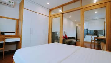 A bedroom with a bed, a desk, and a wardrobe. There is a kitchenette in the background with a refrigerator and a microwave.