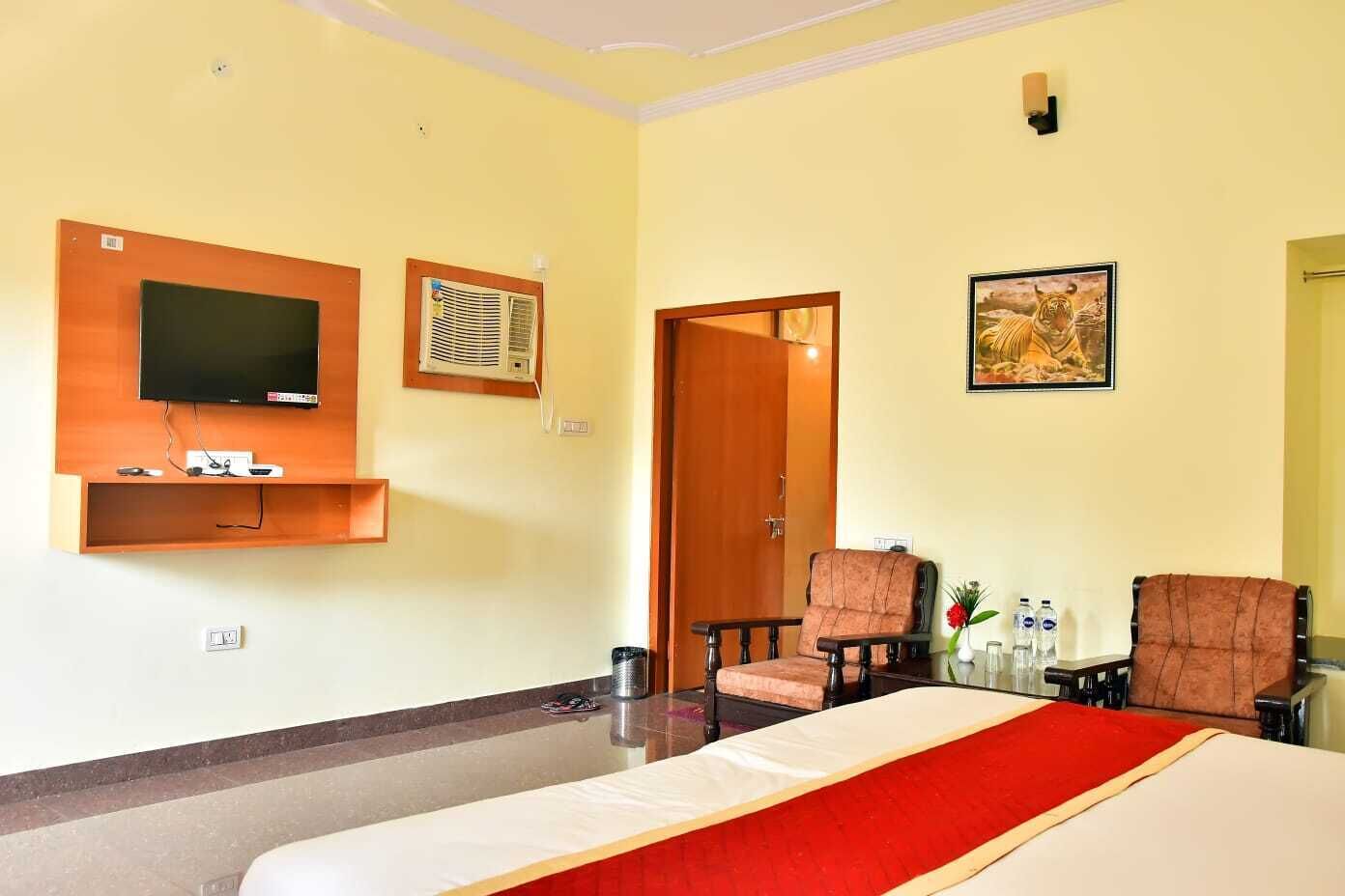 Une chambre d’hôtel avec un lit, une télévision, un appareil de climatisation, deux chaises, une porte et un tableau encadré au mur.