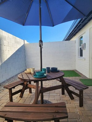 Eine Terrasse mit einem Holztisch und Bänken, einem blauen Sonnenschirm und einer weißen Backsteinmauer.