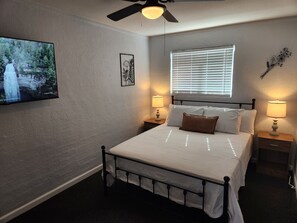 Ein Schlafzimmer mit einem Bett, zwei Nachttischen, einem Deckenventilator, einem Fenster mit Jalousien und einem an der Wand montierten Fernseher.