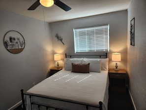 Ein Schlafzimmer mit einem Bett, zwei Nachttischen mit Lampen, einem Deckenventilator, einem Fenster mit Jalousien und Wandbildern.