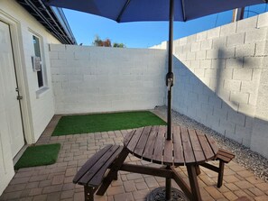 Eine Terrasse mit Holztisch und Bänken, einem blauen Sonnenschirm und einer weißen Ziegelwand.