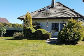 Ein Haus mit Ziegeldach, Schornstein und einer Haustür mit Glasverzierungen. Vor dem Haus befinden sich gepflegte Büsche und eine Rasenfläche.