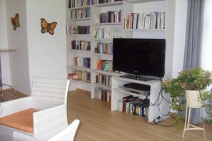 A living room with a bookshelf, a flat-screen TV, a white wicker chair, and a potted plant. - Brünnstein holiday apartment complex (Oberaudorf)