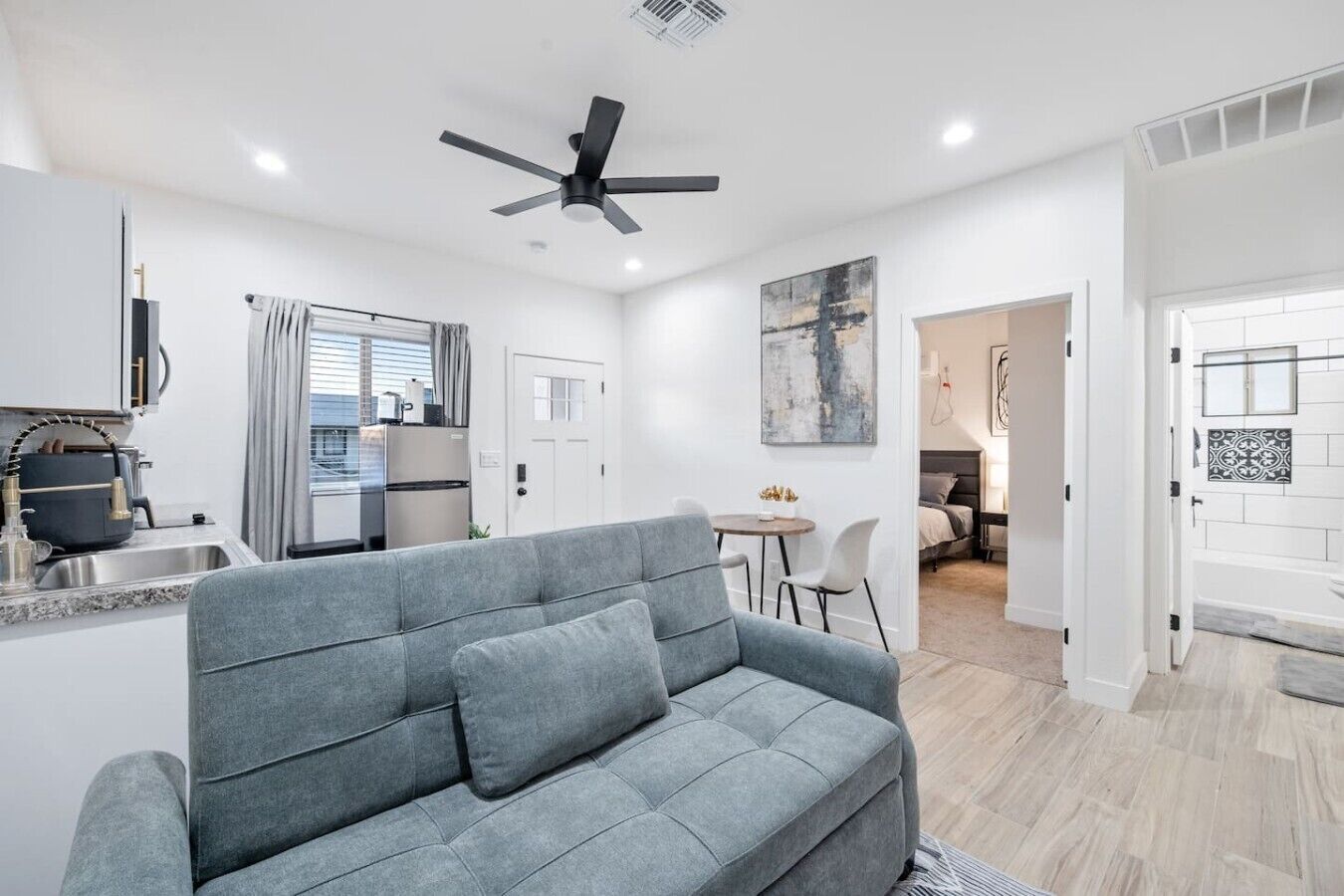 A modern living space with a grey sofa, a dining area, a kitchenette, and a bedroom.