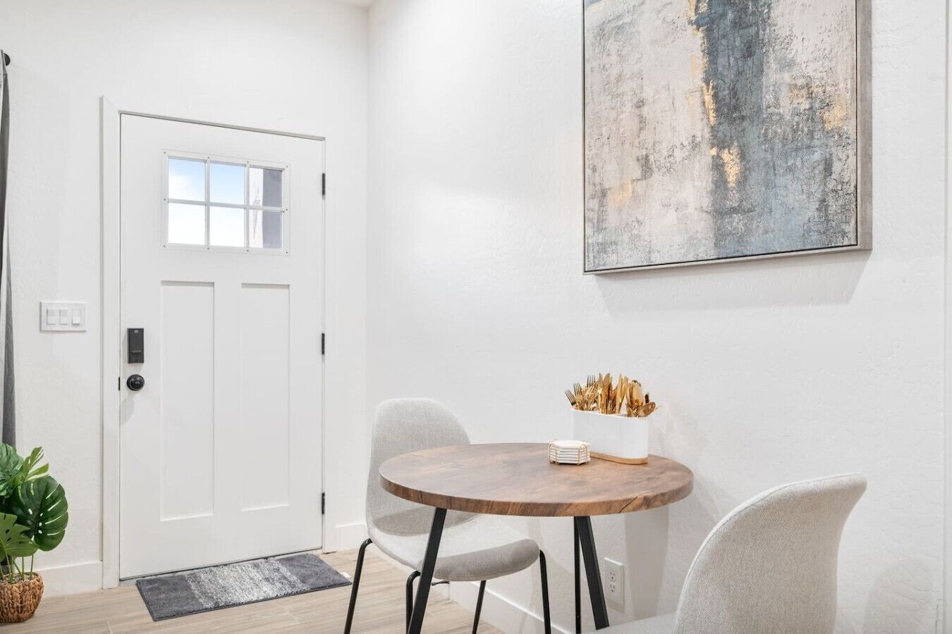 Une pièce avec une porte blanche, une table ronde en bois avec deux chaises, un vase blanc contenant des ustensiles et un grand tableau abstrait accroché au mur.