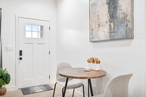 A room with a white door, a round wooden table with two chairs, a white vase with utensils, and a large abstract painting on the wall. - Chic 1-Bedroom Retreat - Phoenix (Phoenix)