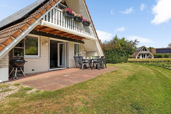 A house with a sloped roof, a balcony with hanging flower boxes, a barbecue grill, and a patio with outdoor furniture. - Holiday apartment with 3 bedrooms (Gramsbergen)