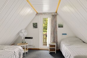 A small, cozy attic bedroom with two beds, a fan, a small table, and a window with curtains. - Holiday apartment with 3 bedrooms (Gramsbergen)