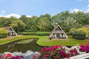 A tranquil lakeside setting with two cottages, each featuring a balcony and a small deck, surrounded by lush greenery and a pond with lily pads. - Holiday apartment with 3 bedrooms (Gramsbergen)