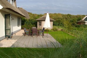 Une terrasse avec une table et des chaises, un parasol blanc et un bâtiment à toit de chaume.