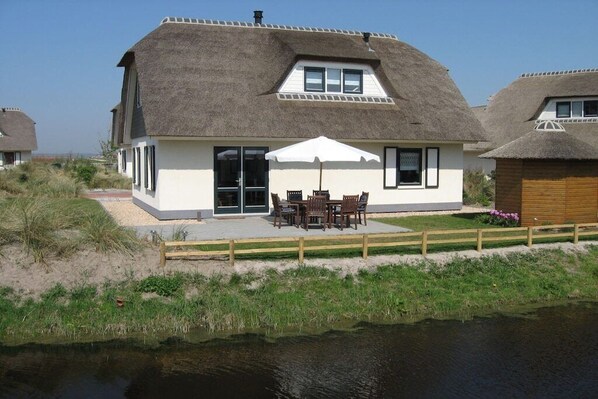 Une maison avec un toit de chaume, une terrasse avec des meubles d’extérieur et une vue sur un plan d’eau.