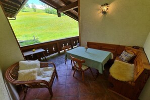 A covered patio with a table and chairs, a wicker chair with a white cushion, a wooden bench with a fur throw, and a view of a green field. - Lauchlehen - Fewo Göll (Bischofswiesen)