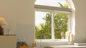 A modern kitchen with a white countertop, a sink, and a window with a view of trees. There is a bowl of fruit on the counter and a lamp on the left.
