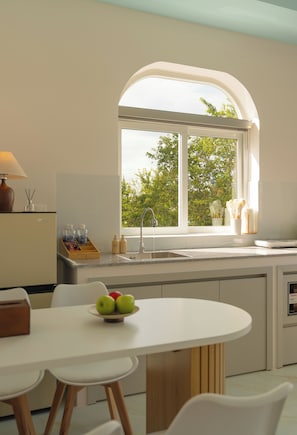 Een moderne keuken met een wit aanrecht, een spoelbak en een raam met uitzicht op bomen. Op het aanrecht staat een schaal met fruit en een lamp aan de linkerkant.