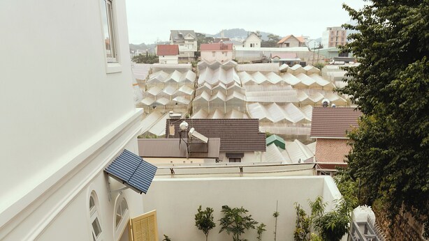Uma área de refeições ao ar livre com uma mesa comprida posta para seis pessoas, rodeada por cadeiras brancas, num pátio com vista para edifícios vizinhos.